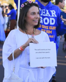 Woman dressed in white and smiling holding a sign that reads "The awful words you hear are called hate speech. I am not with this man." In front of a man wearing a shirt that reads "Fear God" and who is holding a bullhorn.